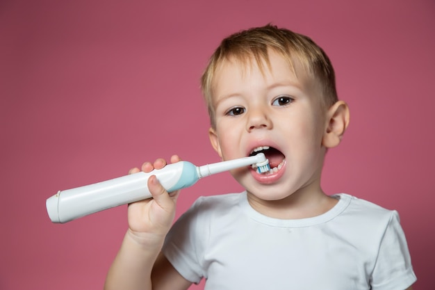 Sonriente niño caucásico limpiando sus dientes con cepillo de dientes sónico eléctrico sobre fondo rosa.