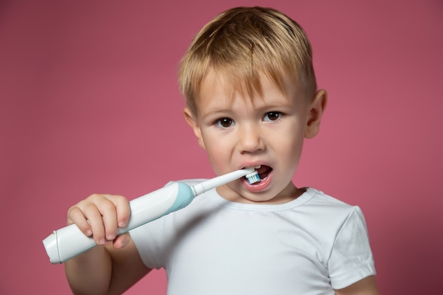 Foto sonriente niño caucásico limpiando sus dientes con cepillo de dientes sónico eléctrico sobre fondo rosa.