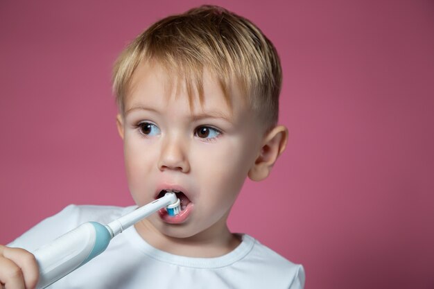 Sonriente niño caucásico limpiando sus dientes con cepillo de dientes sónico eléctrico sobre fondo rosa.