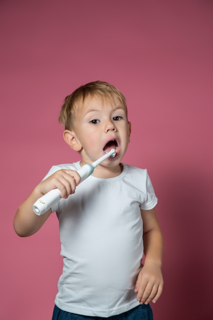 Foto sonriente niño caucásico limpiando sus dientes con cepillo de dientes sónico eléctrico sobre fondo rosa.