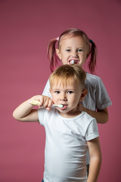 Sonriente niño y niña caucásicos limpiando sus dientes con cepillo de dientes eléctrico sónico y manual sobre fondo rosa.