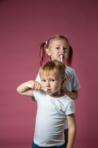 Sonriente niño y niña caucásicos limpiando sus dientes con cepillo de dientes eléctrico sónico y manual sobre fondo rosa.