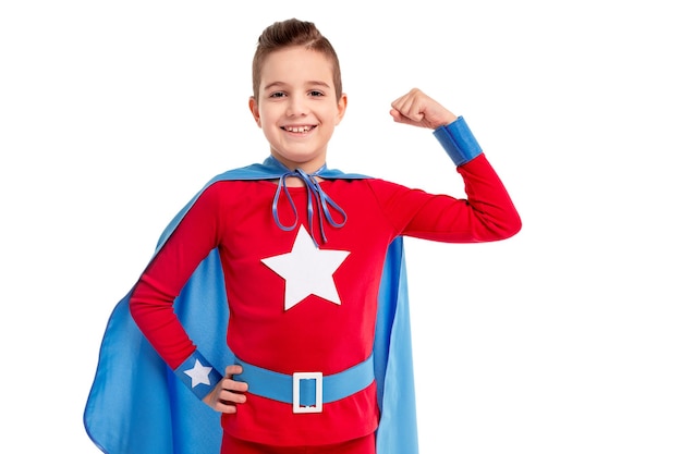 Sonriente niño preadolescente fuerte seguro en traje de superhéroe brillante con capa mostrando bíceps y mirando a cámara contra el fondo blanco.