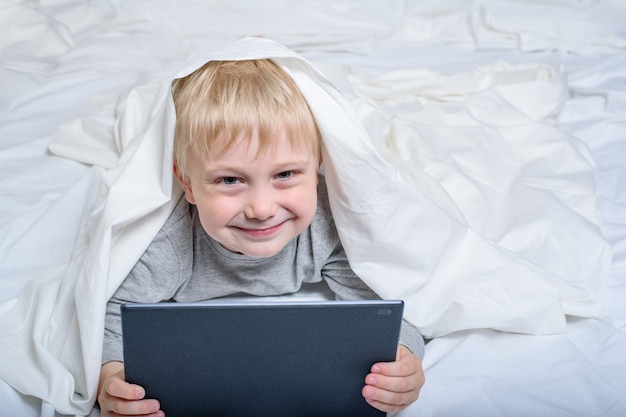 Sonriente niño rubio con una tableta. Acostado en la cama y escondido debajo de las sábanas. Ocio Gadget