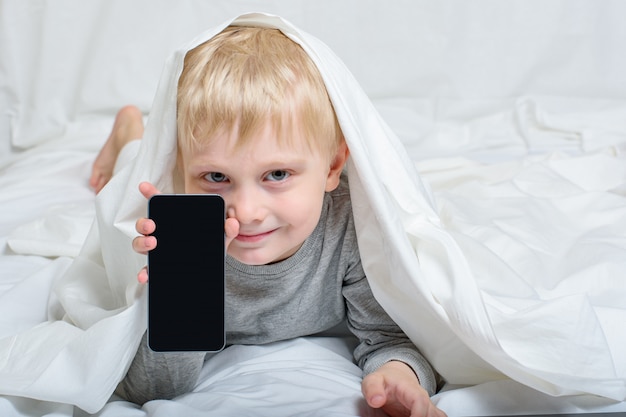 Sonriente niño rubio con un teléfono inteligente. Acostado en la cama y escondido debajo de las sábanas. Ocio Gadget