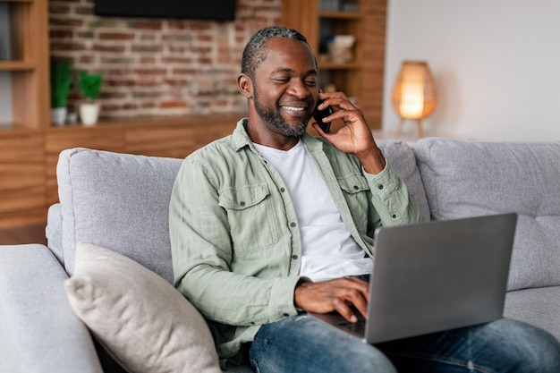 Sonriente ocupado hombre de negocios afroamericano de mediana edad en llamadas casuales por teléfono y escribiendo en la computadora portátil