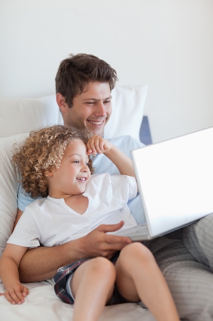 Sonriente padre e hijo en la cama con la computadora portátil