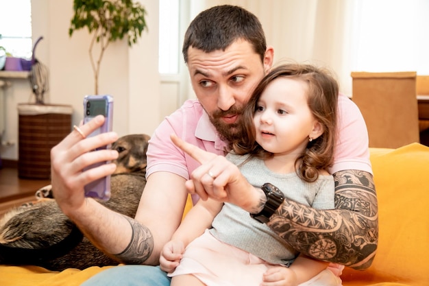 Sonriente padre feliz con su pequeña hija linda mirando la pantalla del teléfono inteligente tomando una fotografía de retrato selfie sentado en el sofá divirtiéndose juntos concepto de familia feliz