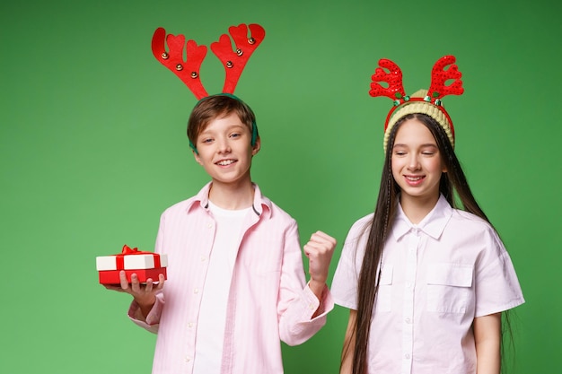 Sonriente pareja de adolescentes caucásicos en traje de navidad con cuernos de venado en la cabeza sosteniendo una caja de regalo ...