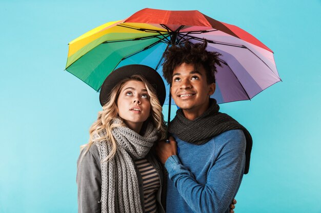 Foto sonriente pareja de adolescentes multirraciales vistiendo ropa de invierno de pie bajo un paraguas aislado sobre la pared azul