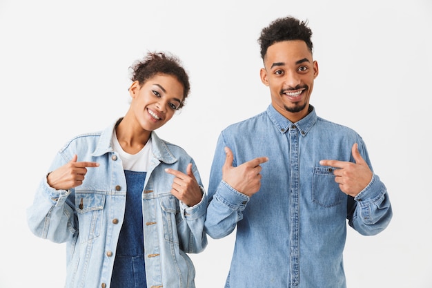 Sonriente pareja africana en camisas de mezclilla apuntando sobre sí mismo y sobre la pared gris