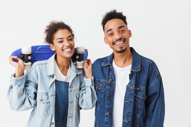 Sonriente pareja africana en camisas de mezclilla posando juntos mientras la mujer sostiene patineta sobre pared gris