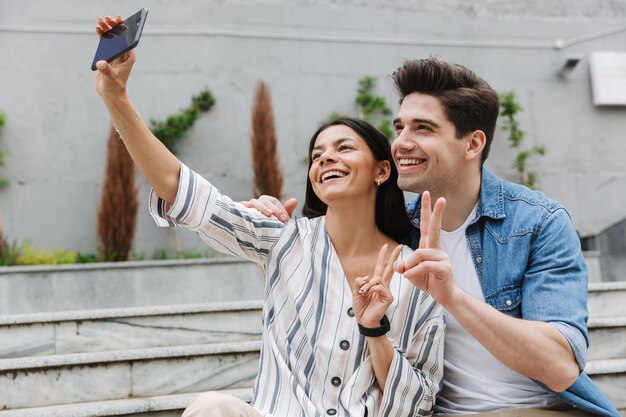 Sonriente pareja amorosa joven optimista positiva al aire libre tomar un selfie por teléfono móvil mostrando paz.