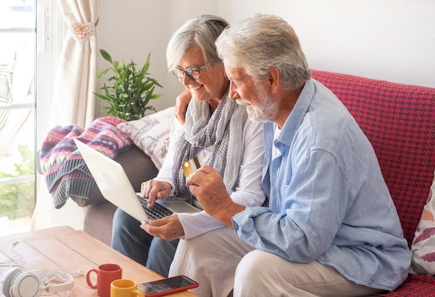 Sonriente pareja de ancianos comprando en línea con una laptop sentada en un sofá en casa Viejas y atractivas personas de cabello gris pagan con el concepto de consumismo bancario con tarjeta de crédito