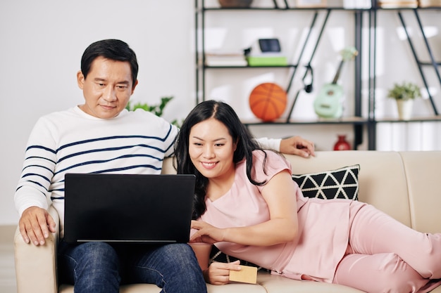 Sonriente pareja asiática de mediana edad relajándose en casa y haciendo compras en línea a través de una computadora portátil