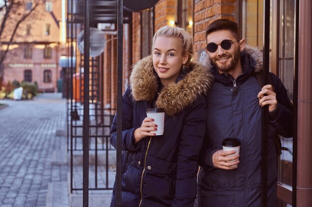 Sonriente pareja atractiva con abrigos de invierno sosteniendo un café para llevar mientras está de pie cerca del café afuera.