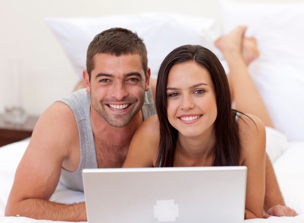 Sonriente pareja en la cama usando una computadora portátil