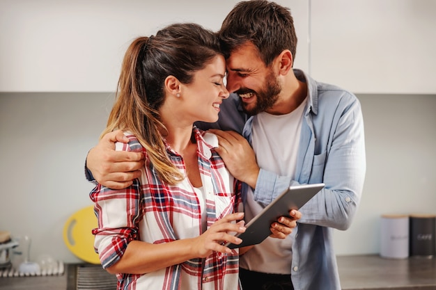 Sonriente pareja feliz de pie juntos en la cocina y usando la tableta para buscar la receta.