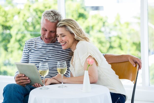 Sonriente pareja madura con tableta digital con vino en la mesa