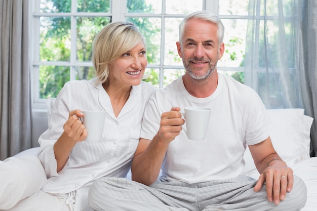 Sonriente pareja madura con tazas de café sentado en la cama