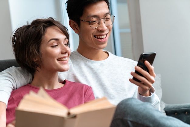 Sonriente pareja multinacional leyendo un libro y usando el teléfono celular mientras está sentado en el sofá en casa