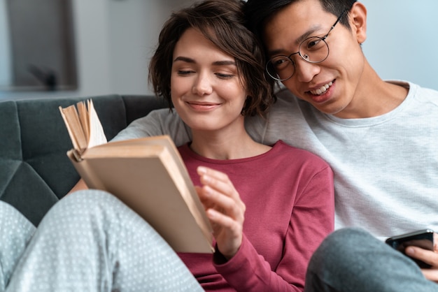 Foto sonriente pareja multinacional leyendo un libro y usando el teléfono celular mientras está sentado en el sofá en casa