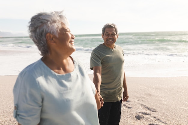 Sonriente pareja multirracial senior cogidos de la mano mientras disfruta de un día soleado en la orilla de la playa