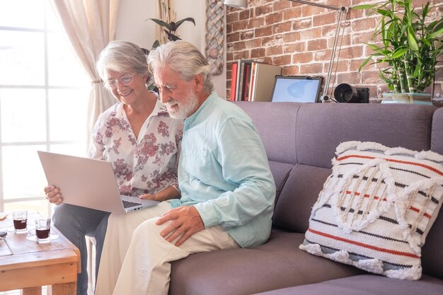 Sonriente pareja senior en el sofá en casa usando la computadora portátil con un descanso con café expreso