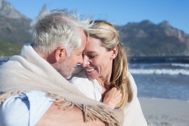 Sonriente pareja sentada en la playa bajo una manta