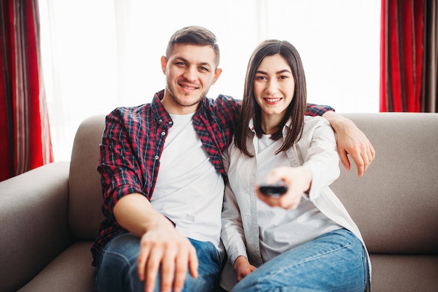 Sonriente pareja sentada en el sofá y ver la televisión en casa, mujer con control remoto en mano