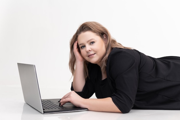 Sonriente y pensativa mujer de cabello rubio acostada en el piso usando una computadora portátil en un fondo blanco independiente