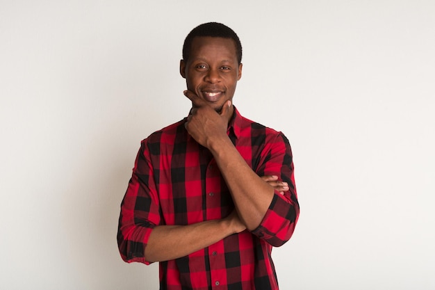 Sonriente, pensativo joven negro en retrato de camisa a cuadros roja en casual en el fondo blanco del estudio. Chico en camisa a cuadros