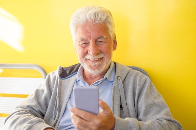 Sonriente y relajado anciano de cabello blanco sentado con fondo amarillo usando teléfono móvil