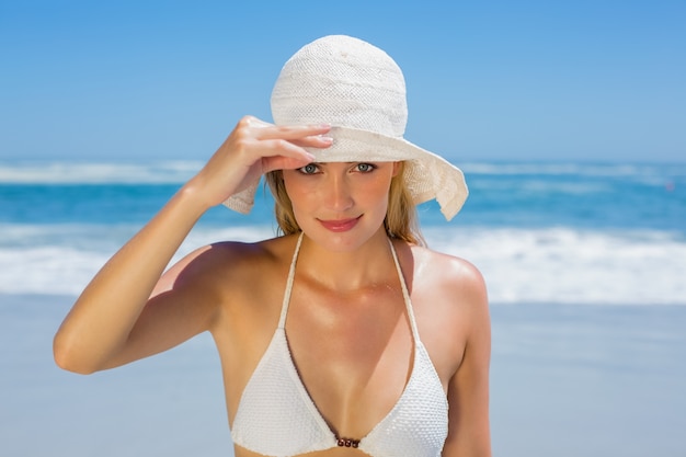 Sonriente rubia en bikini blanco y sombrero para el sol en la playa