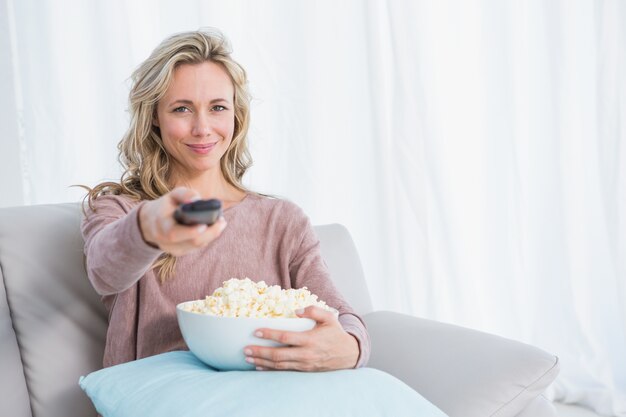 Sonriente rubia cambiando el canal de televisión mientras come palomitas de maíz