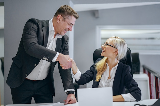 Sonriente rubia empresaria caucásica sentado en la oficina y estrecharme la mano con su director. Todos los que conoces tienen algo que enseñarte.