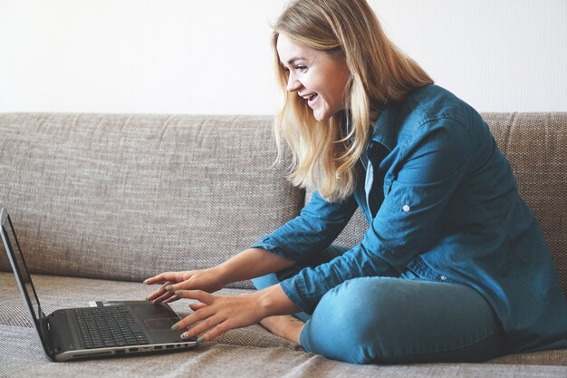 Sonriente rubia feliz relajándose en el sofá con su computadora portátil en casa en la sala de estar, sin etiquetas