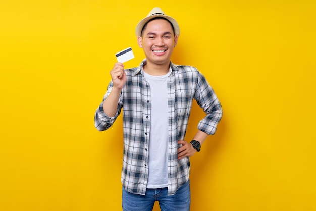 Sonriente satisfecho etnia asiática joven de 20 años con ropa informal sombrero con tarjeta de crédito de pie con la mano en la cintura en el retrato de estudio de fondo amarillo Concepto de estilo de vida de la gente