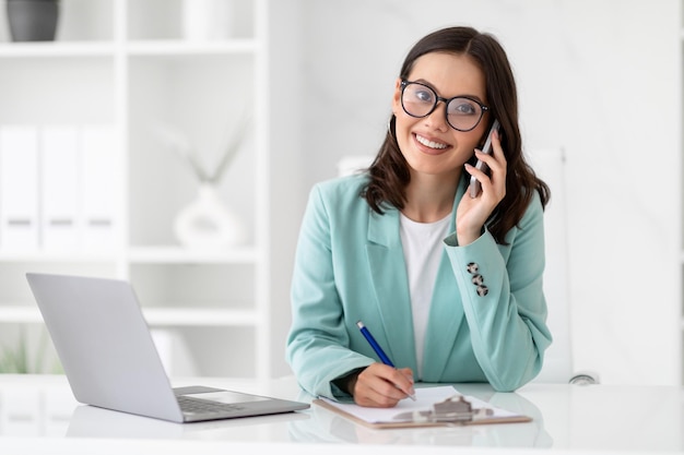 Sonriente secretaria milenaria caucásica con traje y anteojos llama por teléfono en el lugar de trabajo