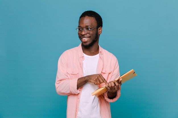 Sonriente sosteniendo carpeta joven chico afroamericano aislado sobre fondo azul.