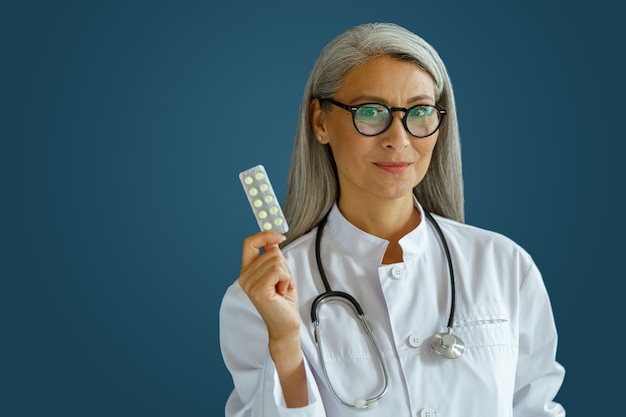 Sonriente terapeuta de mediana edad en uniforme blanco sostiene blister de píldoras de pie sobre fondo azul en estudio. Personal médico profesional