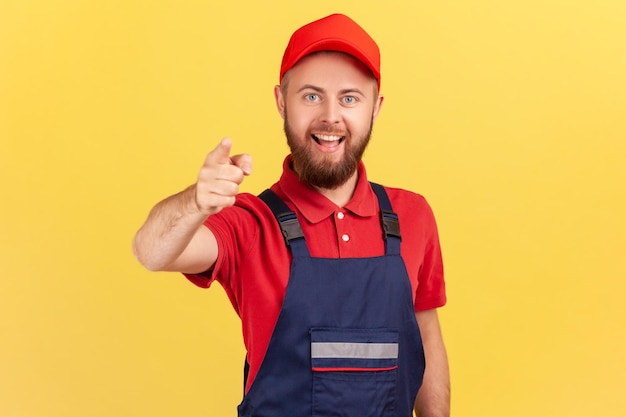 Sonriente trabajador barbudo satisfecho de pie y señalando con el dedo a la cámara eligiéndote