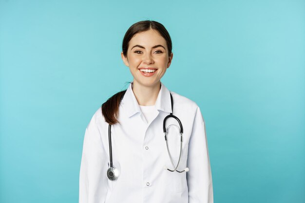 Sonriente trabajadora médica sana, joven doctora que parece feliz, parada en bata blanca y estetoscopio sobre fondo azul.