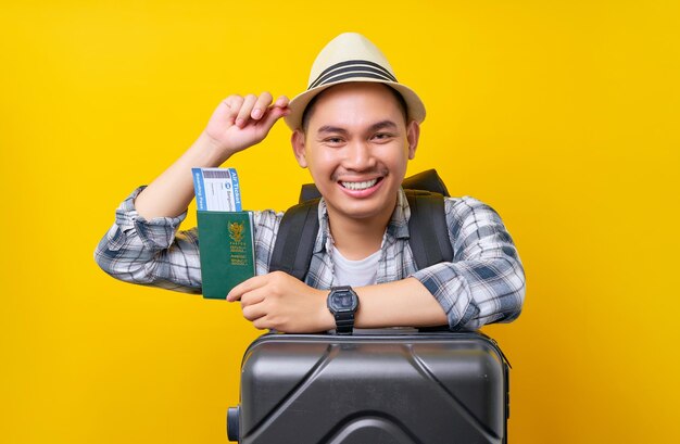 Sonriente turista asiático con ropa informal, sombrero y mochila con billete de pasaporte apoyado en una maleta con fondo amarillo Viaje de pasajeros al extranjero escapada de fin de semana Concepto de viaje en avión