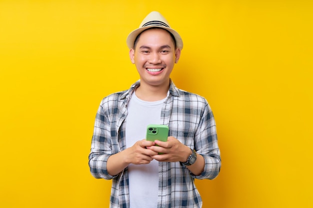 Sonriente viajero turista hombre asiático en ropa casual sombrero sosteniendo teléfono móvil aislado sobre fondo amarillo Pasajero que viaja los fines de semana Concepto de viaje de vuelo aéreo