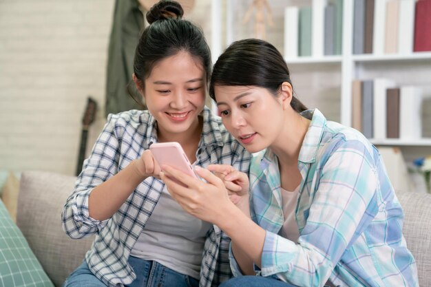 Sonrientes amigas jóvenes disfrutando viendo videos móviles en el teléfono móvil sentados en el sofá en casa. una adolescente positiva sostiene un teléfono inteligente que muestra una foto divertida en línea para que su hermana pase el fin de semana juntos