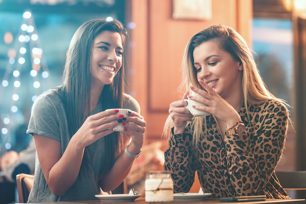 Sonrientes jóvenes queridas amigas están sentadas en el café, hablando y bebiendo café.