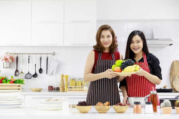 Sonrientes mujeres étnicas en delantales que sirven frutas maduras frescas en la mesa de la cocina.