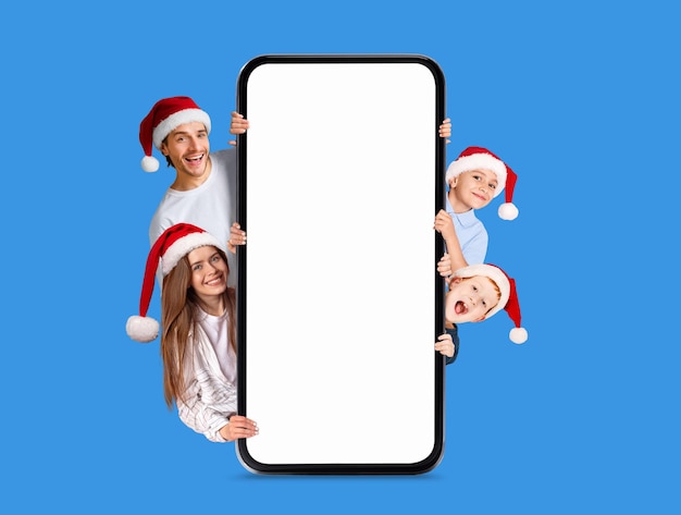 Sonrientes padres jóvenes europeos con niños con sombreros de Navidad asomándose desde un gran smartphone con pantalla vacía