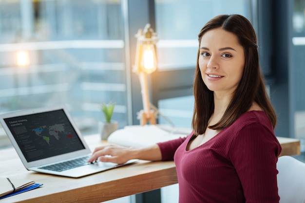 Sonrisa agradable. Cerca de la joven mujer encantadora que le mira mientras usa una computadora portátil y se sienta en la mesa